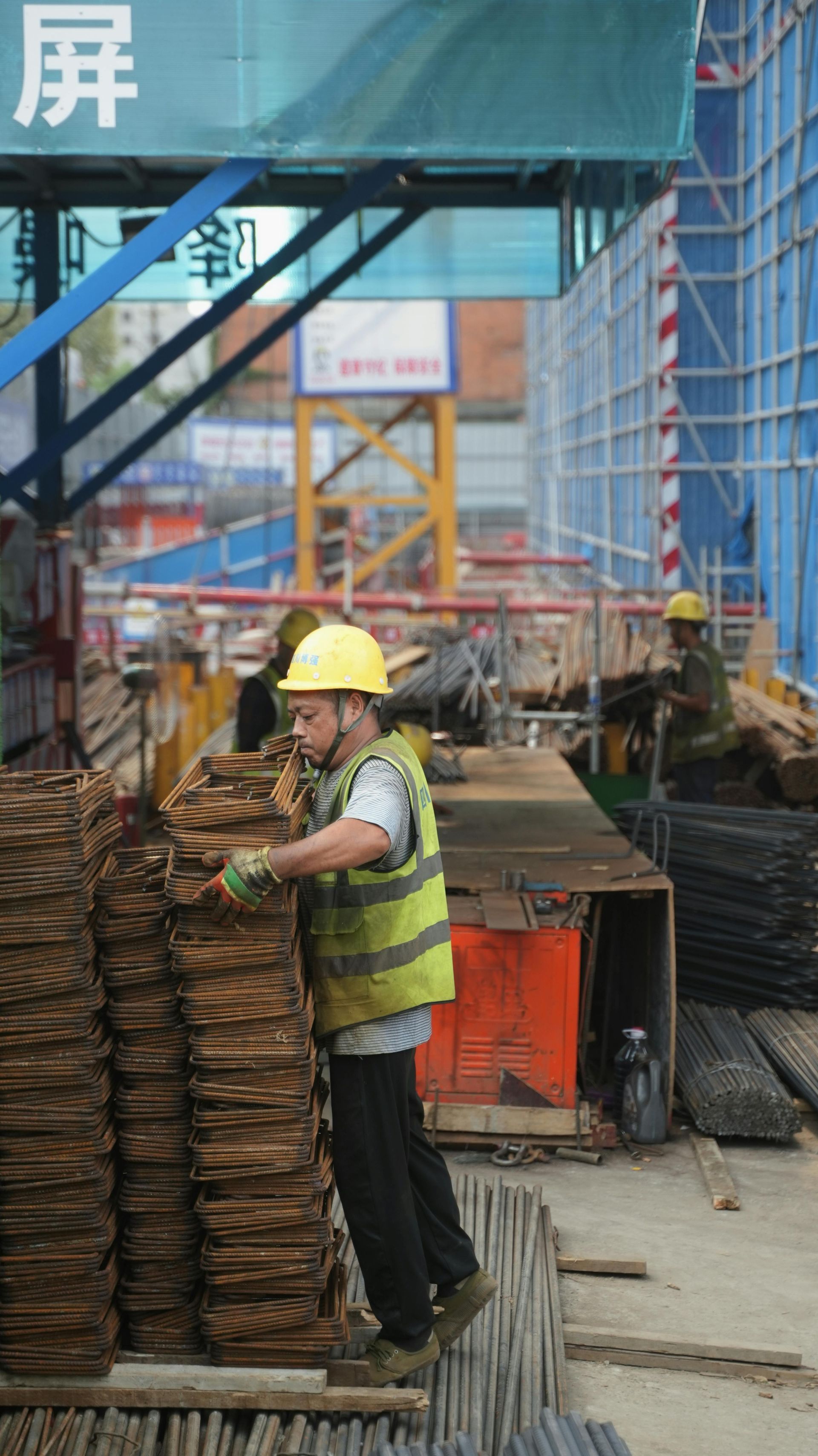 Construction worker wearing yellow hard hat and vest stacks rebar at a building site.