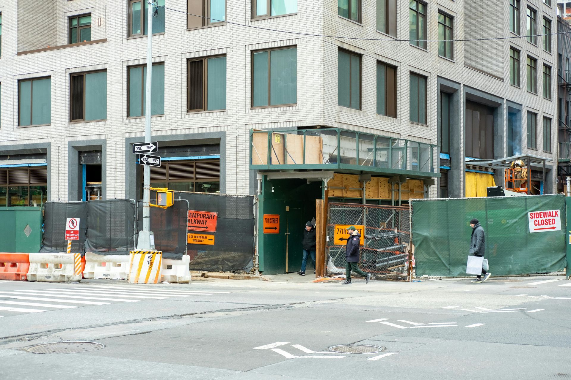 Construction site on a city corner; green fencing, plywood, people walking.