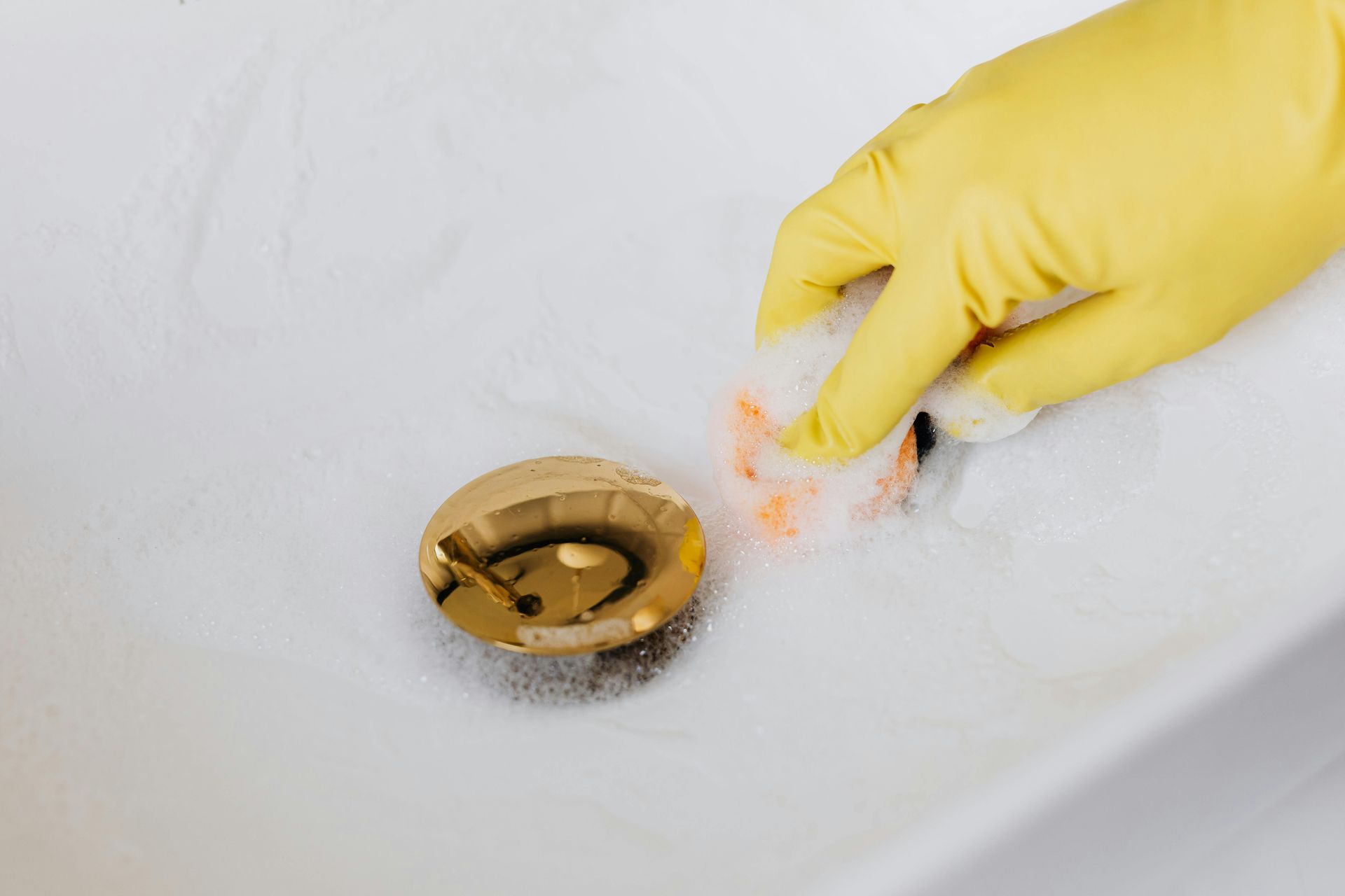 Hand in yellow glove cleaning a white sink with a gold drain.