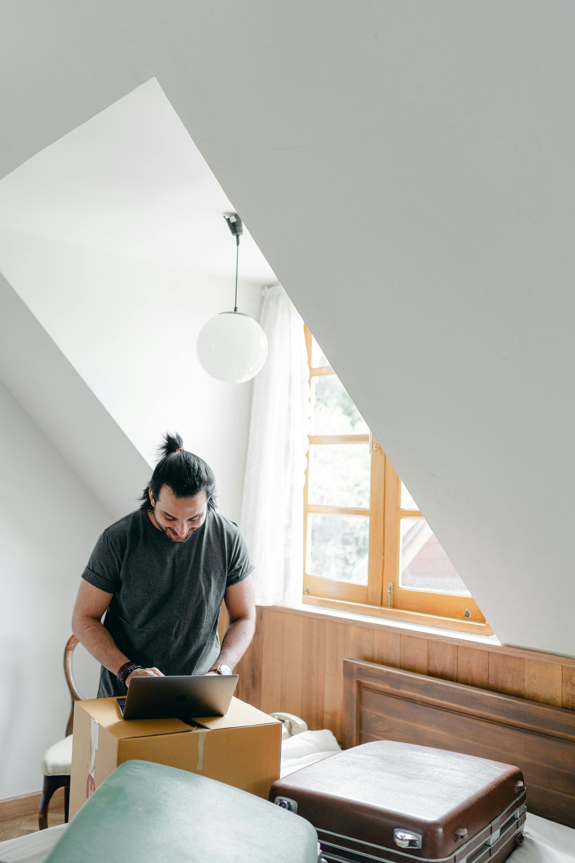 Man in a gray shirt working on a laptop, near boxes and a suitcase in a light-filled room.
