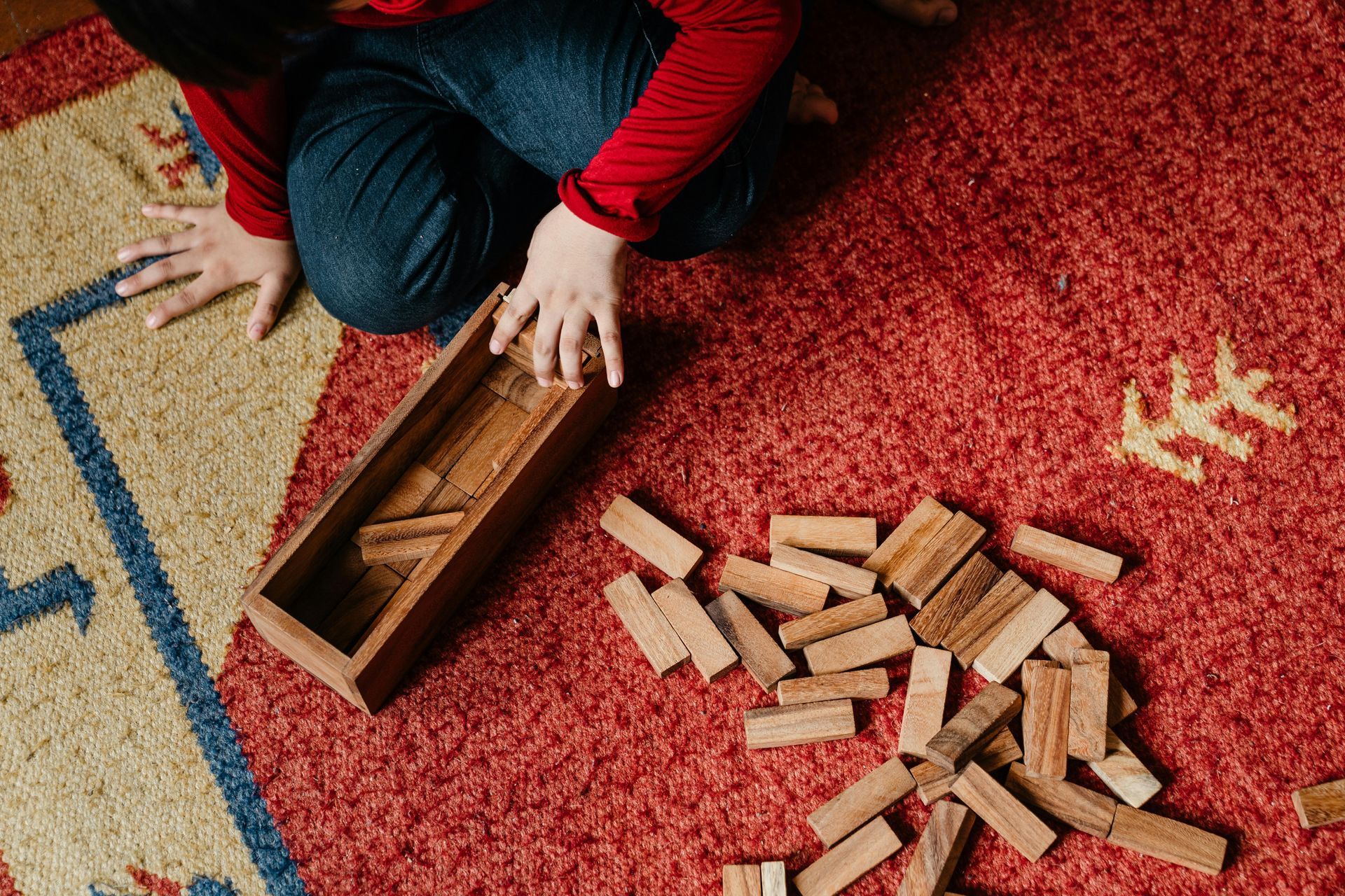 A person kneeling on a red rug, playing with wooden blocks.