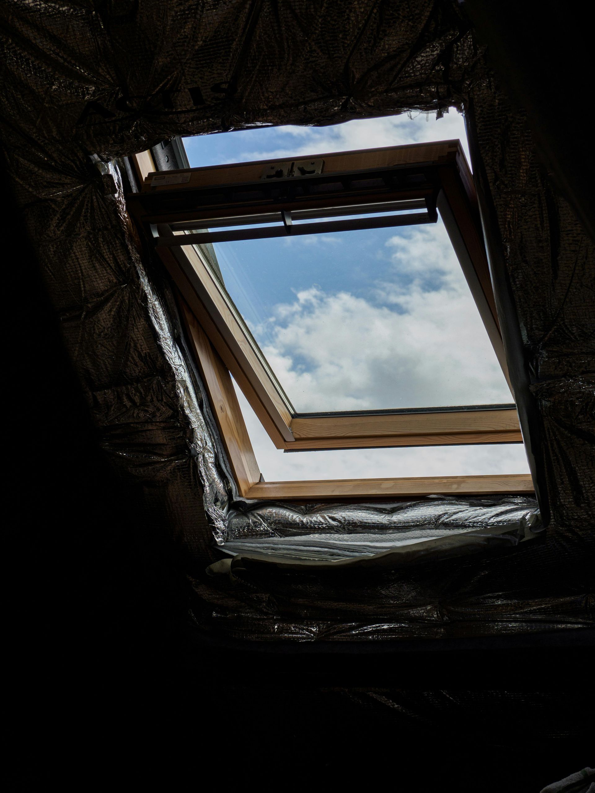 Opened skylight showing a view of the sky with clouds. Wooden frame, dark interior, and reflective insulation.
