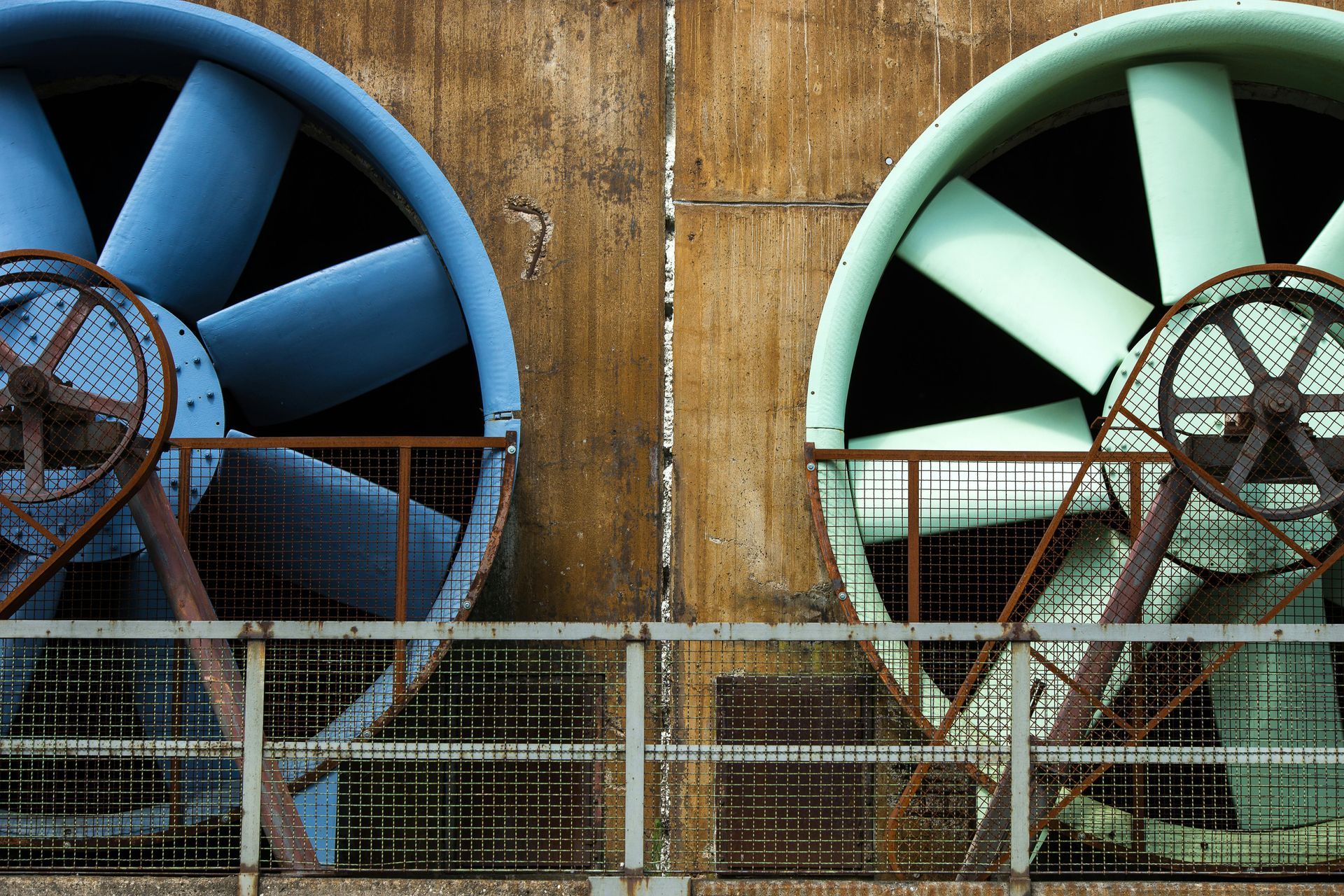 Two large industrial fans, blue and green, against a weathered brown wall.