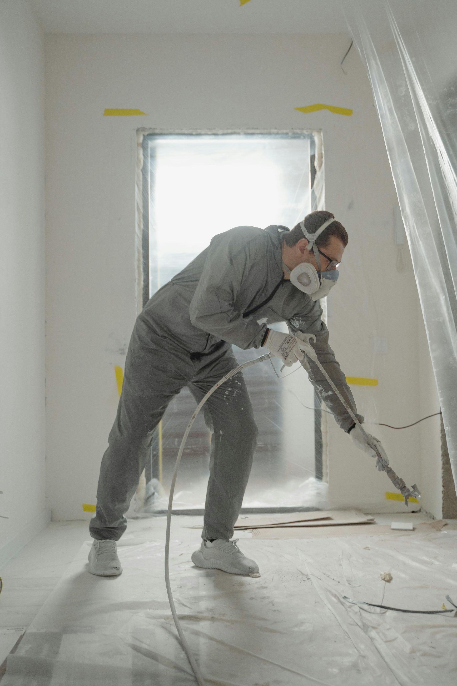 Person in protective suit spraying paint in a room, doorway covered in plastic.
