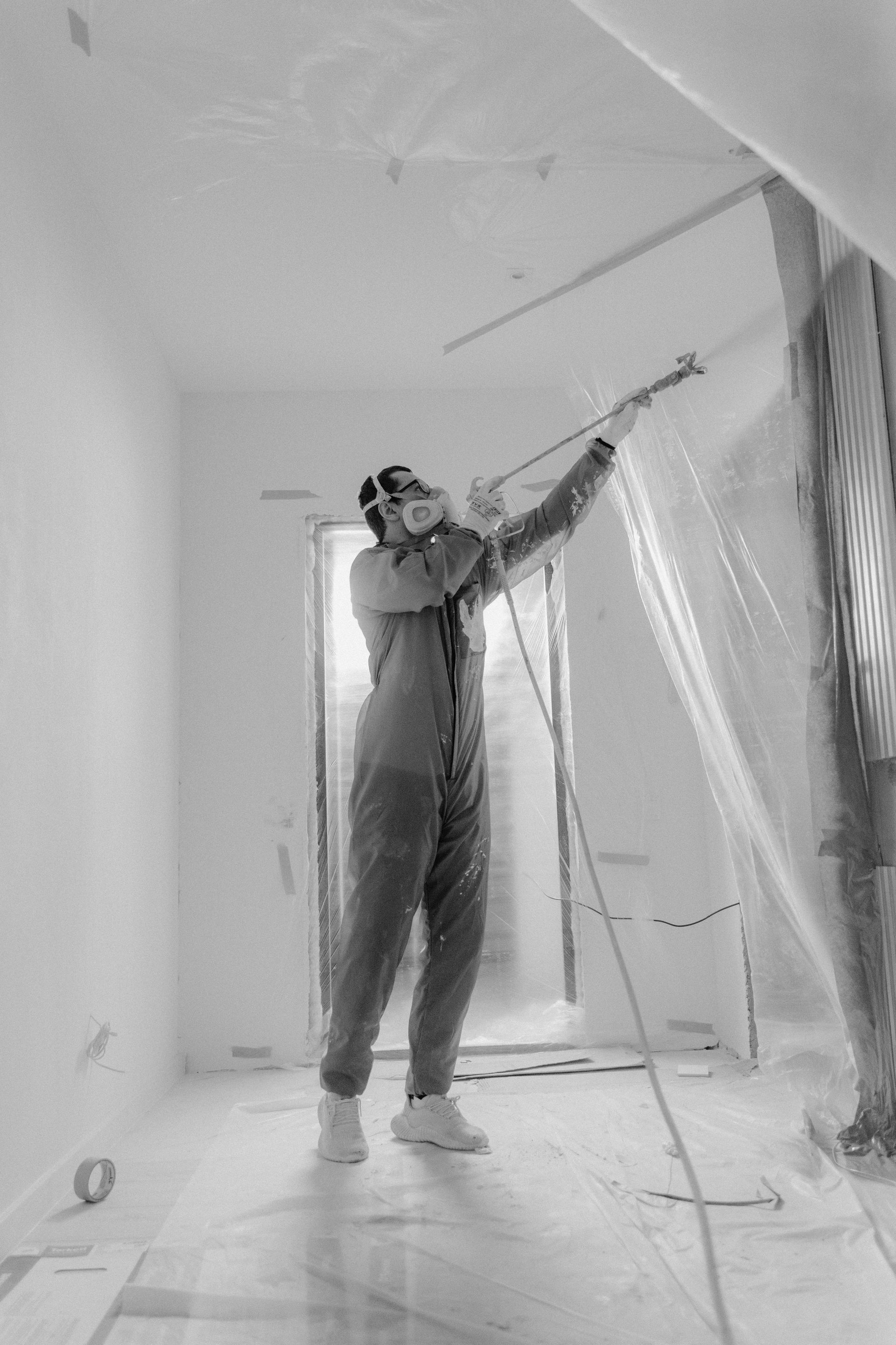 Person in protective suit spraying paint inside a room, with plastic sheeting covering walls and doorway.