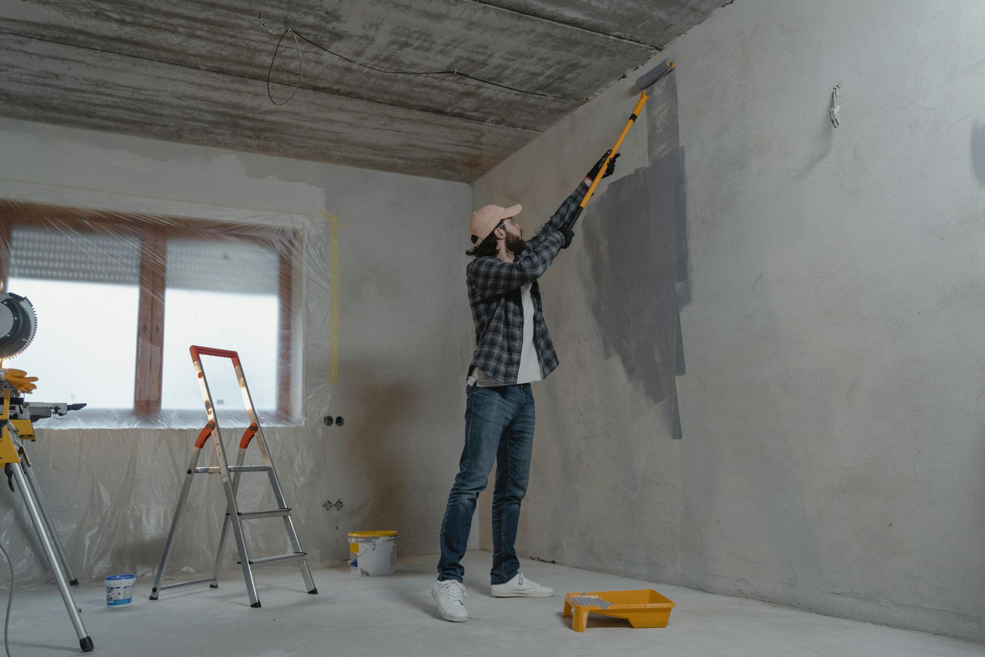Person painting a wall grey with a roller. Interior room with ladder, tools, and window.