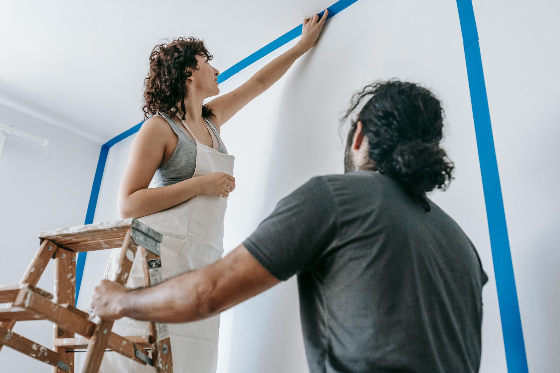 Woman on a ladder taping a wall as a man watches, preparing to paint.