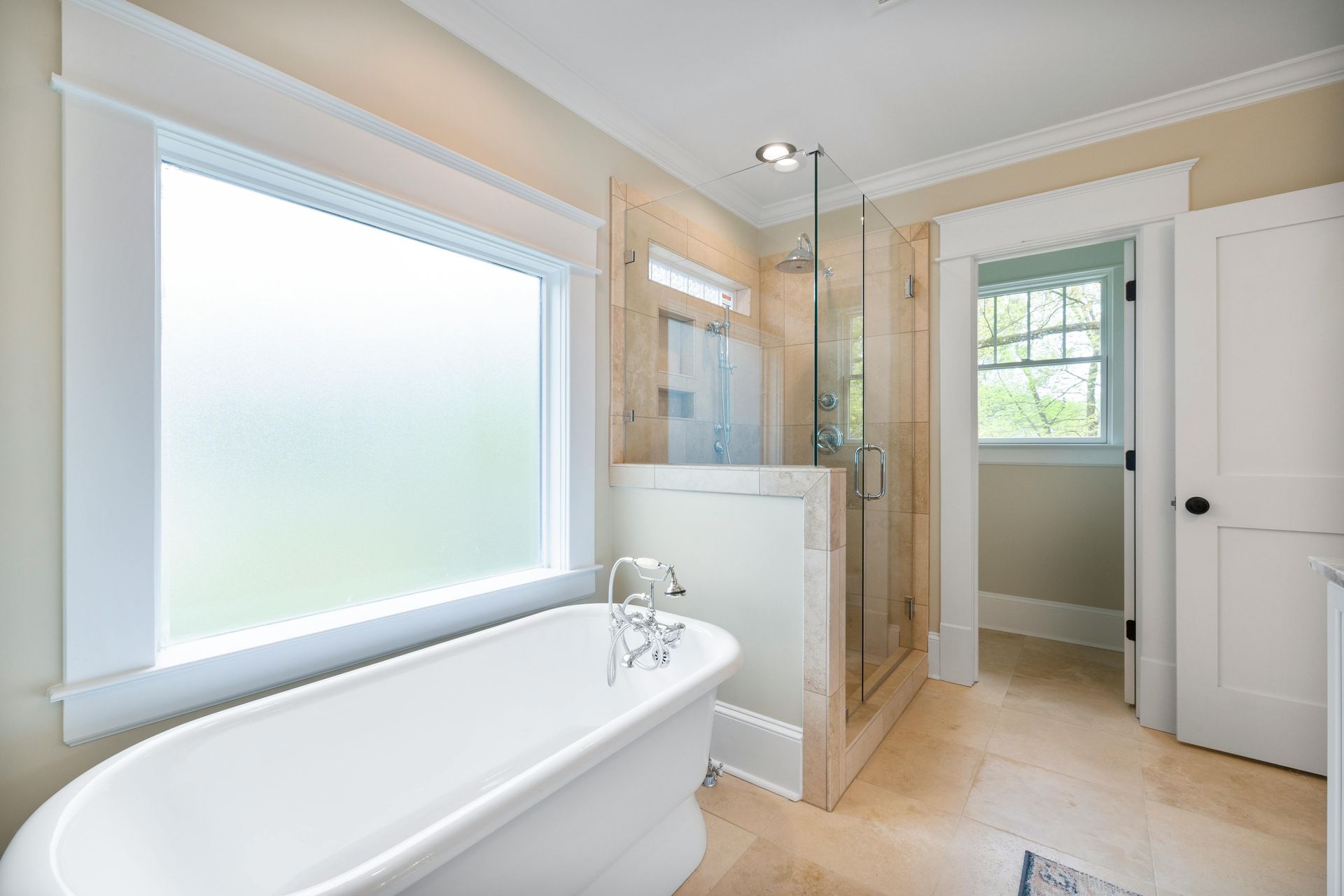 A bright, spacious bathroom with a bathtub, glass shower, and window. Light beige walls and floor.