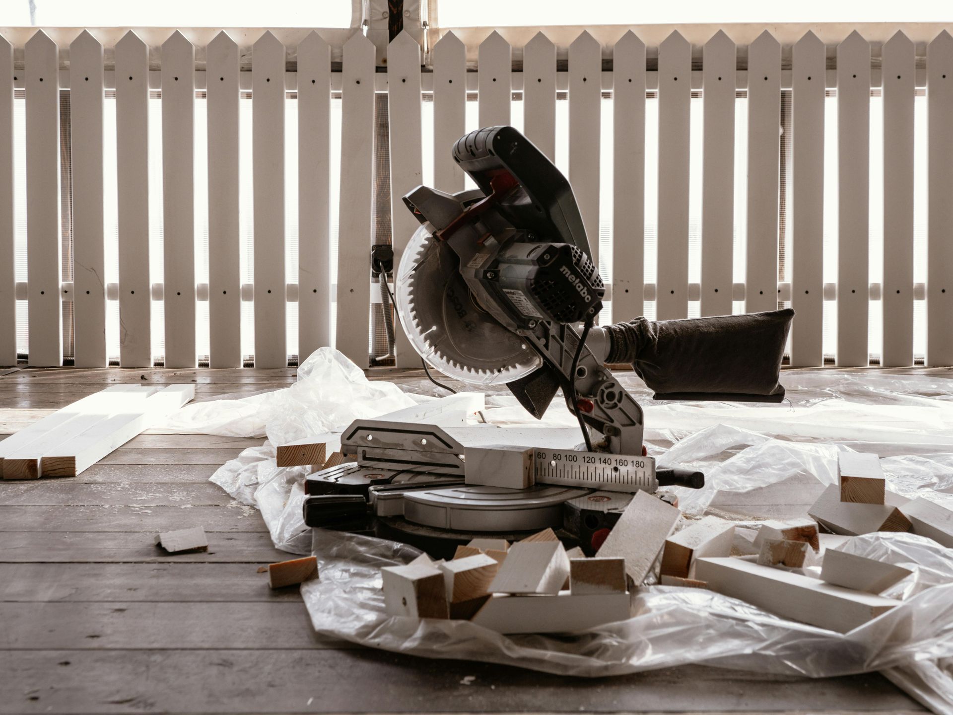 Mitre saw on floor surrounded by wood scraps and plastic sheeting. White picket fence in background.