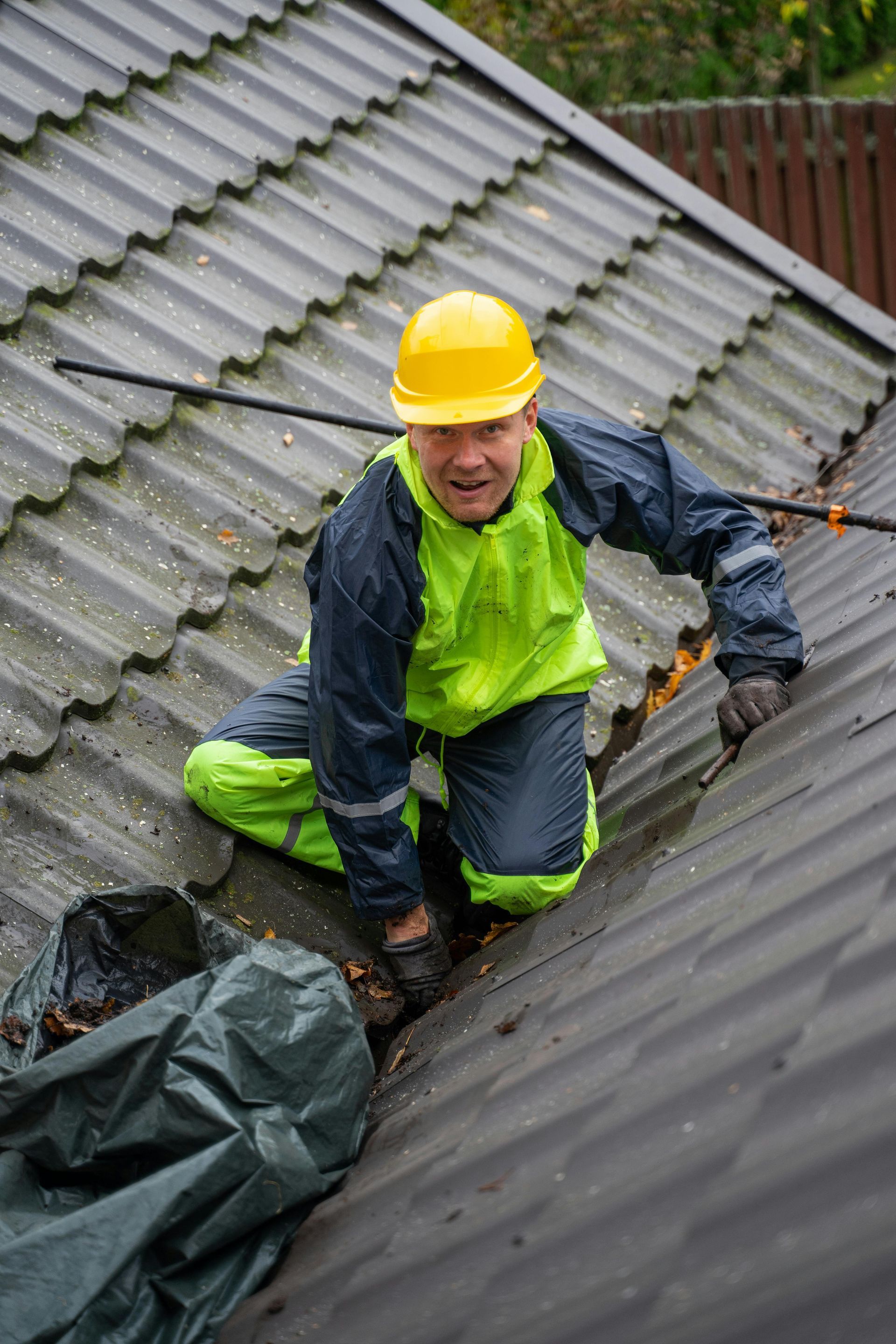 Person on a roof wearing a yellow hard hat and rain gear, cleaning a gutter.