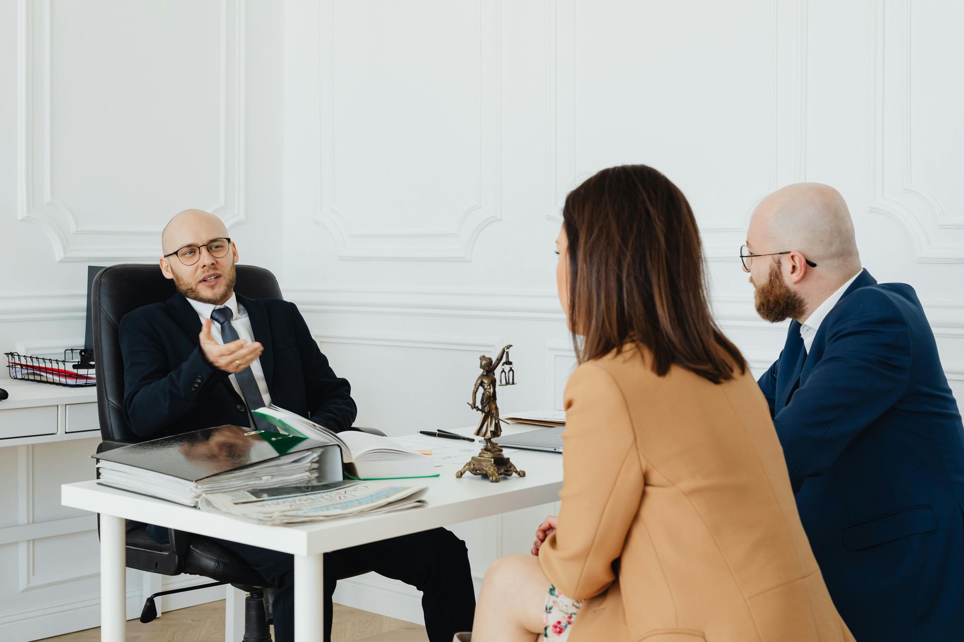 A bald man in a suit speaks at a table to a woman in a tan blazer and a man in a blue suit. Inside.