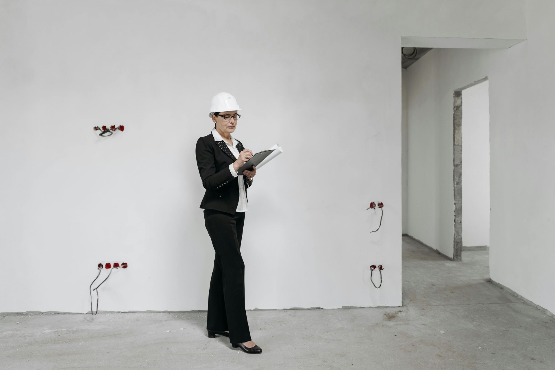 Woman in a hard hat and suit inspecting an unfinished room, taking notes.