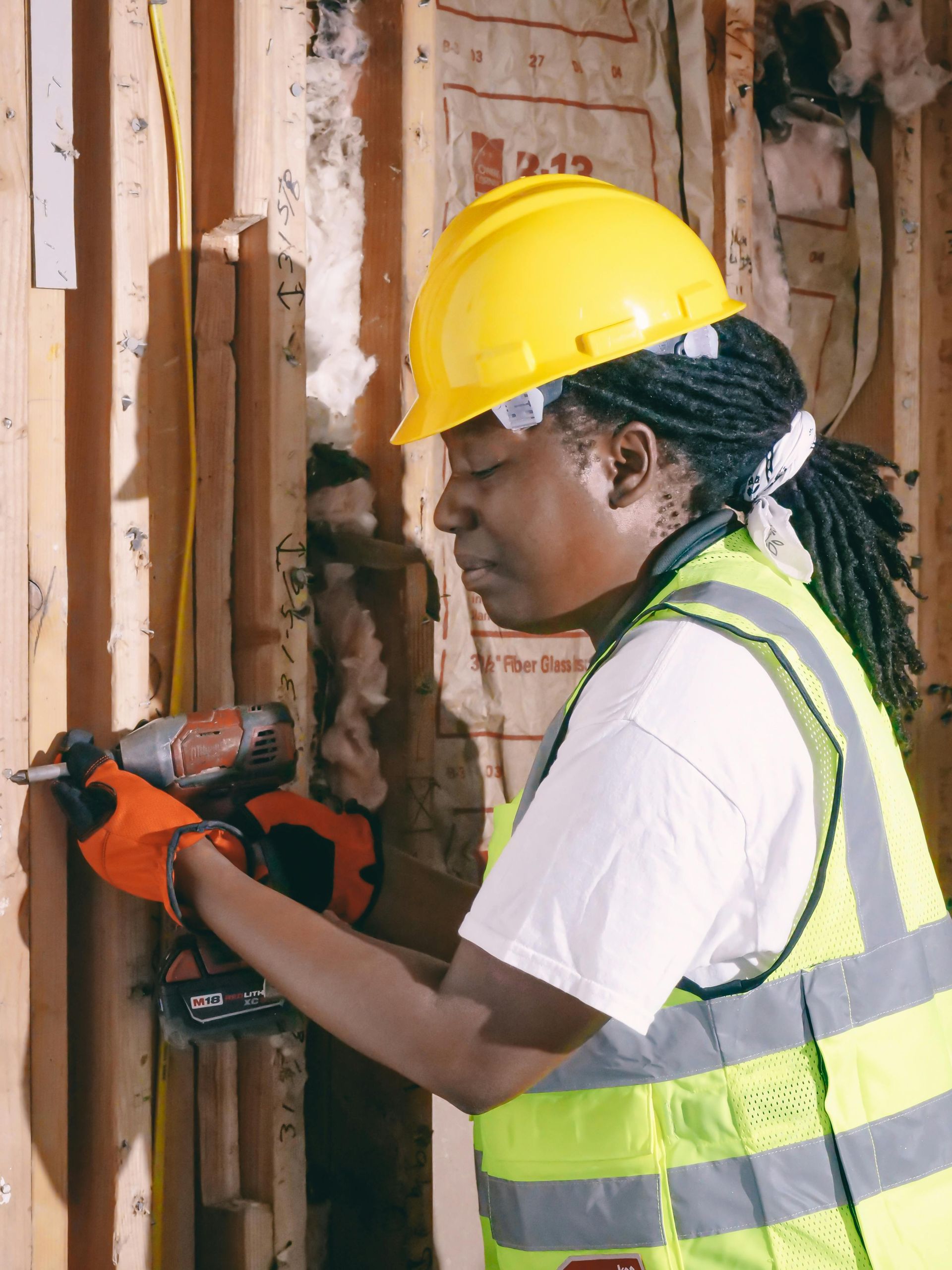 Person in a yellow hard hat and safety vest using a power drill on a wooden wall.