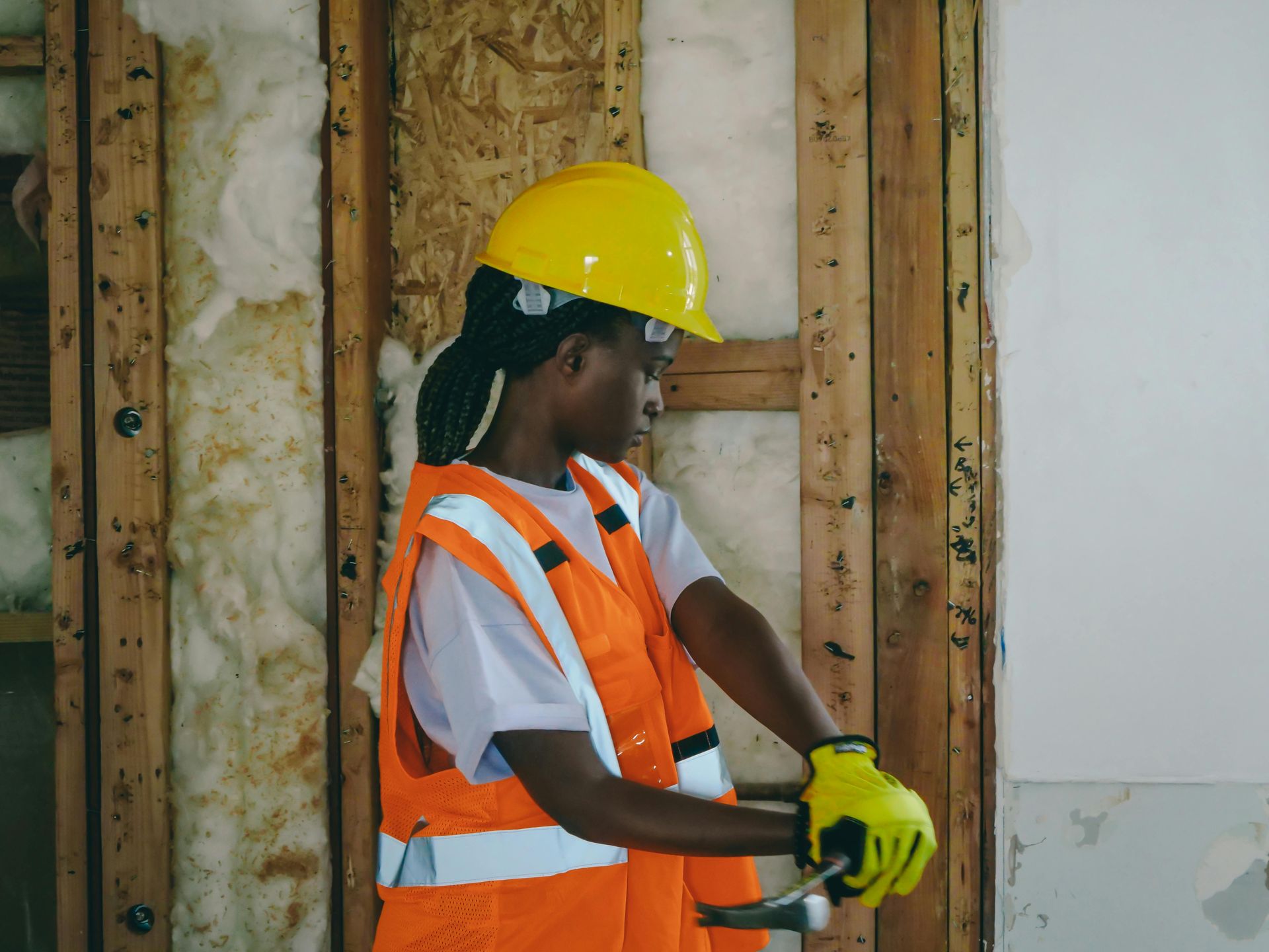Construction worker in yellow hard hat, orange vest, and gloves, hammering on wooden wall.
