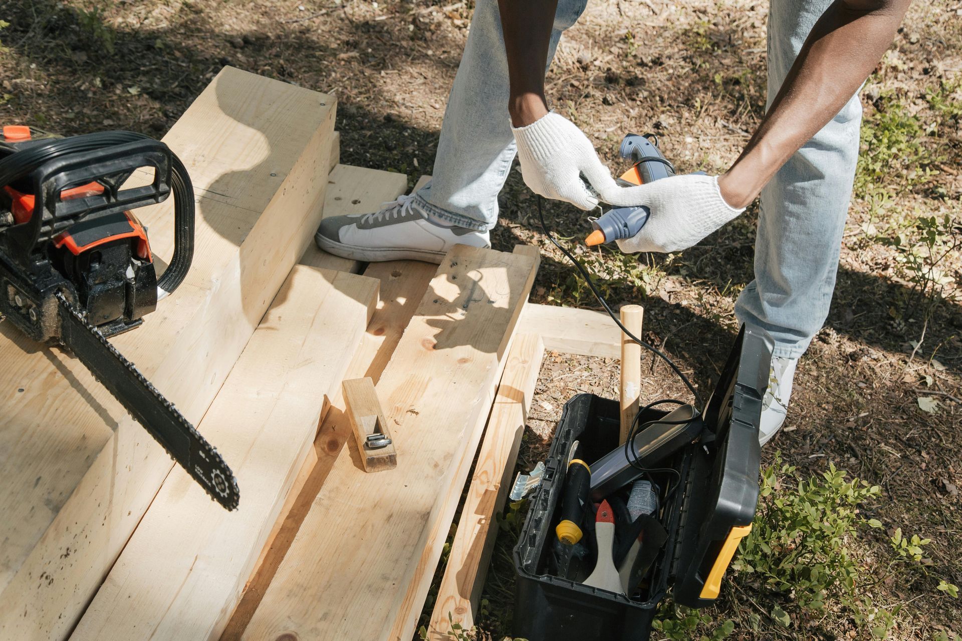 Person wearing gloves and working on wood near chainsaw and toolbox outdoors.