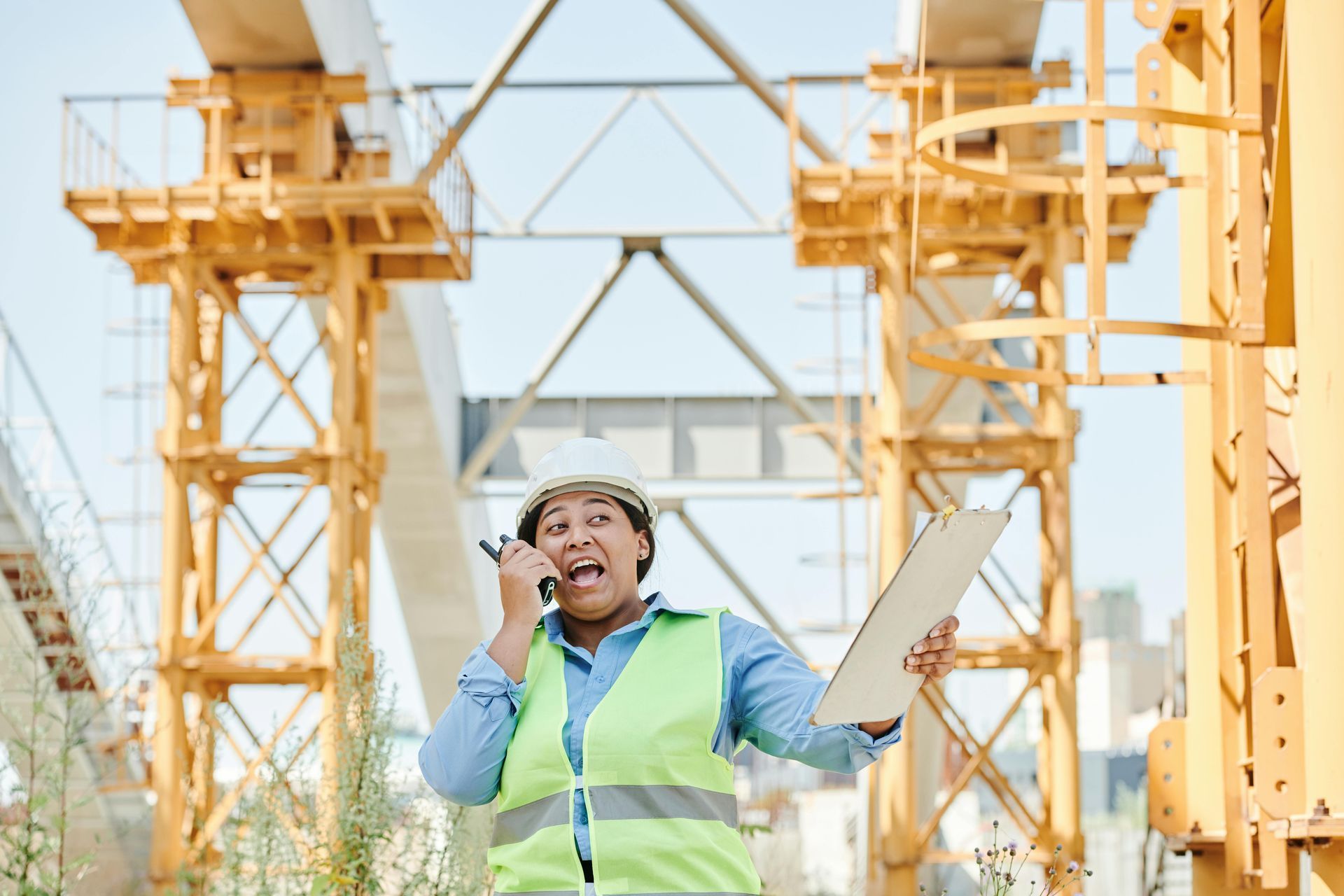 Construction worker, hard hat, and safety vest, talking into a radio on a construction site.