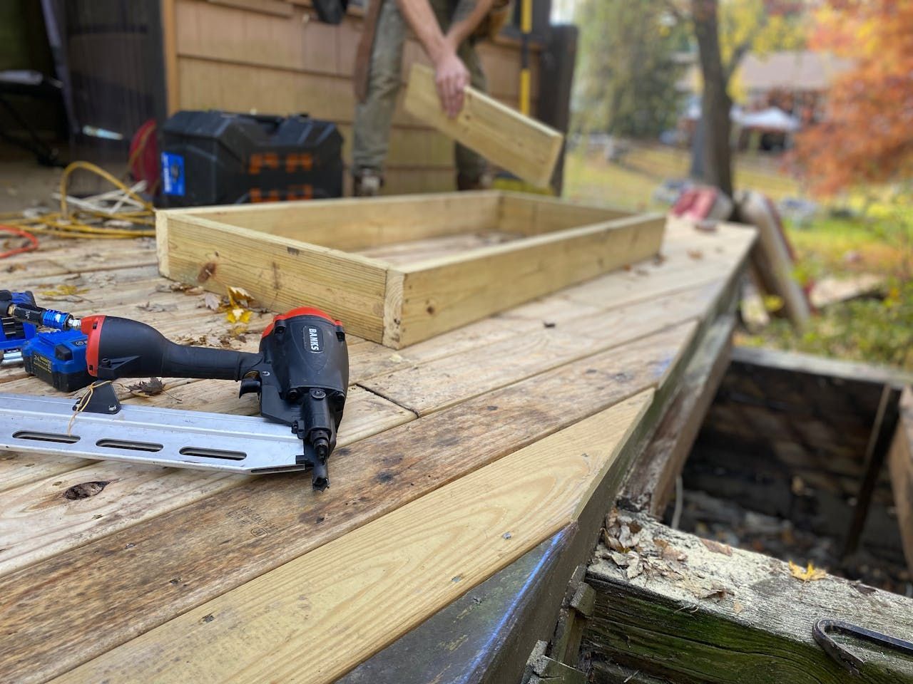 A person building a wooden frame on an outdoor deck, nail gun in foreground.