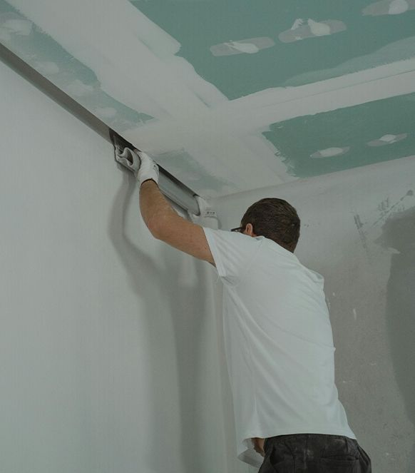 Person applying plaster to a wall with a long trowel in an interior room.