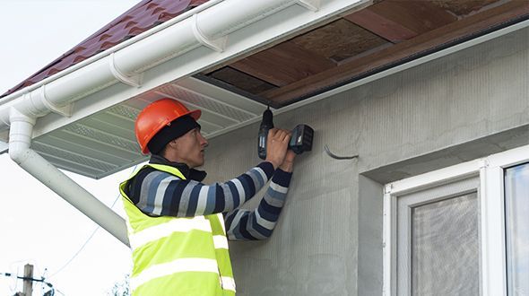Construction worker drills into a house's exterior wall, wearing safety gear near a gutter.