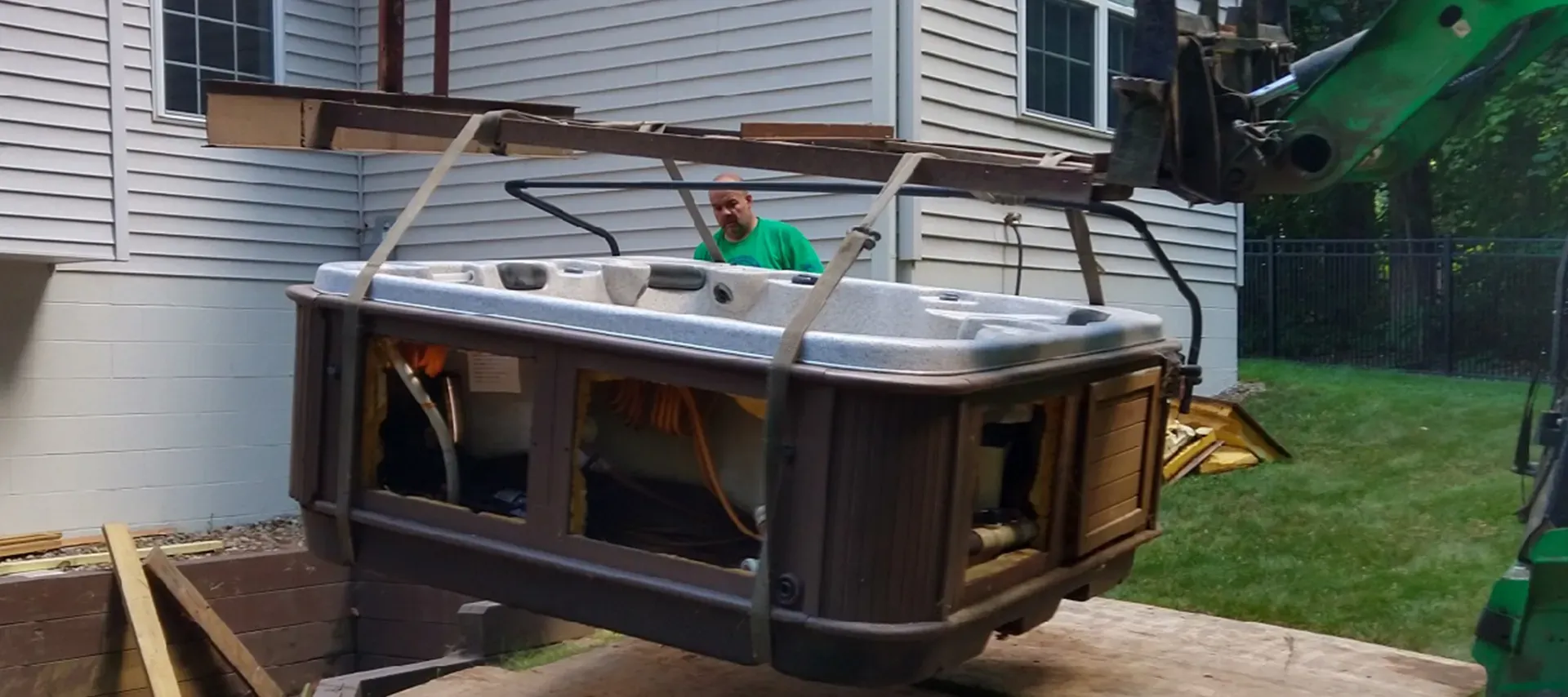 A crane uses straps to lift a large hot tub into place next to a house.