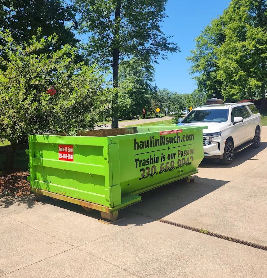 A bright green dumpster for Haulin’n’Such sits on a concrete driveway next to a white SUV on a sunny day.