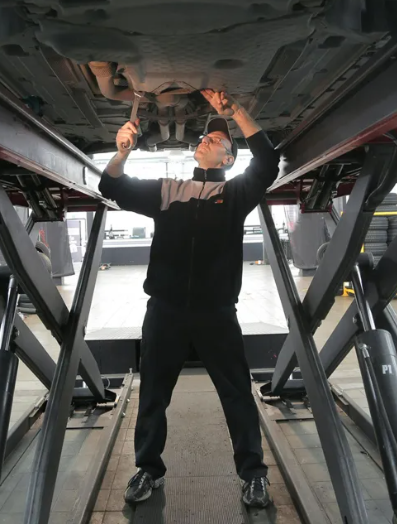 Mechanic under a car on a lift, working with a wrench. Wearing a black jacket and pants. | Bloomfield Garage