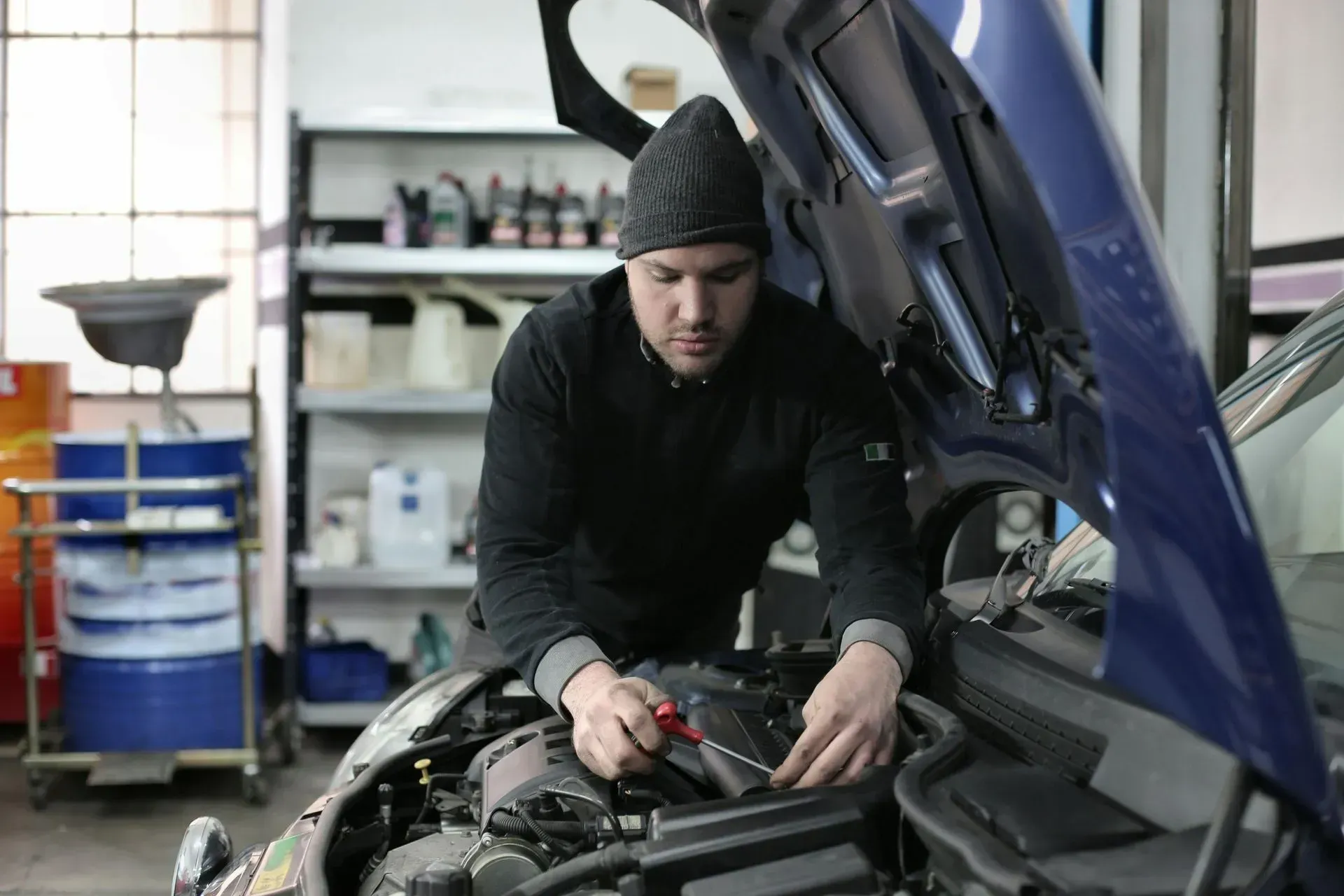 Mechanic in a black hat and sweater working on a car engine in a garage. | Bloomfield Garage