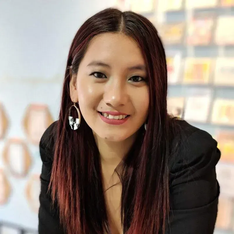 Woman with auburn hair smiles, wearing a black blazer and hoop earrings, in front of a blurred background.