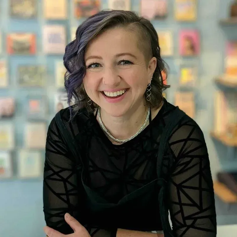 Woman with purple hair smiling, wearing a black top, in front of a wall of cards.