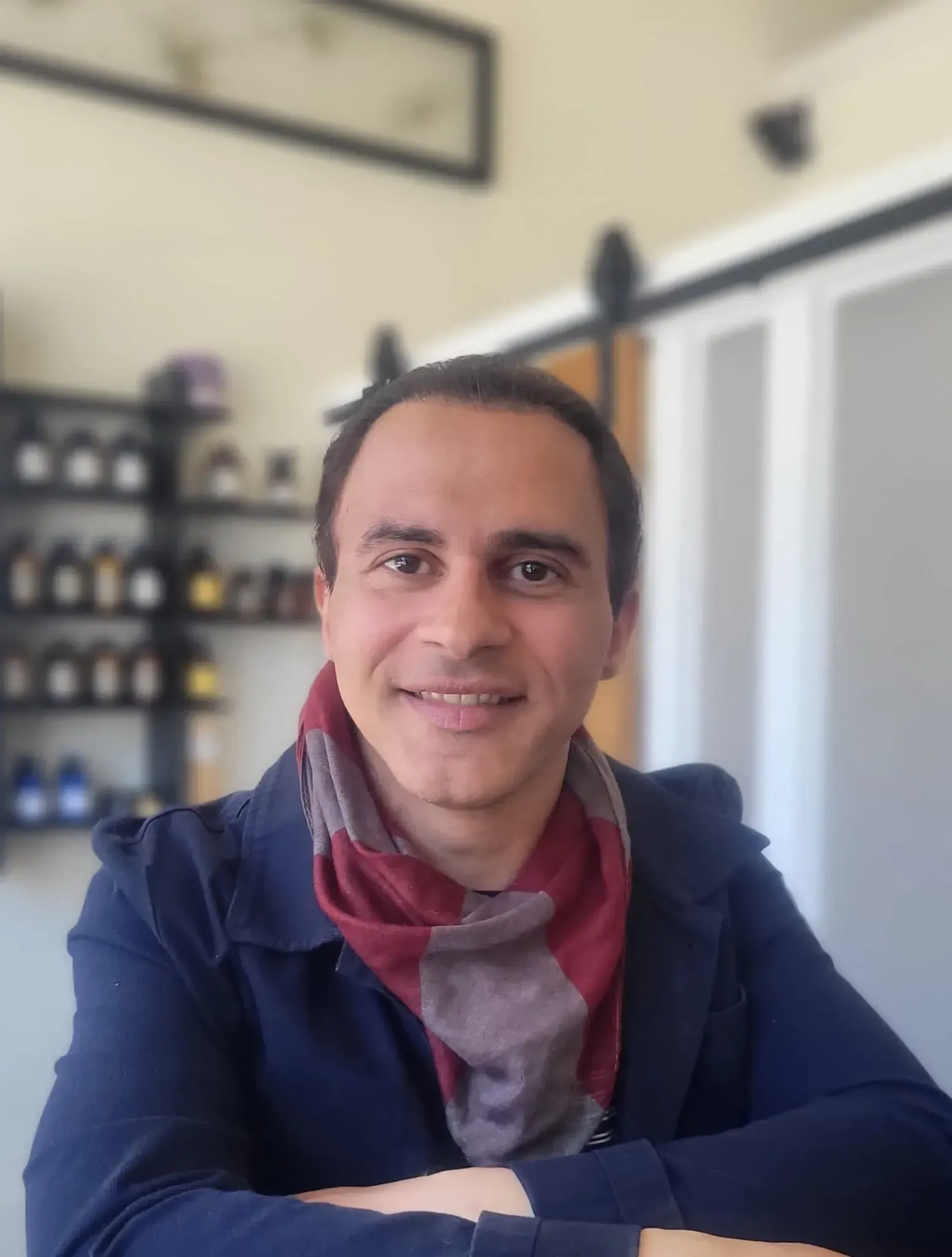 Man smiling, wearing a blue jacket and red scarf, indoors with shelves and a doorway in the background.