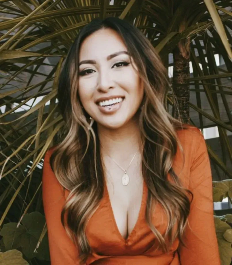 Woman with long, wavy brown hair, wearing a rust-colored dress, smiling at the camera, outdoors.
