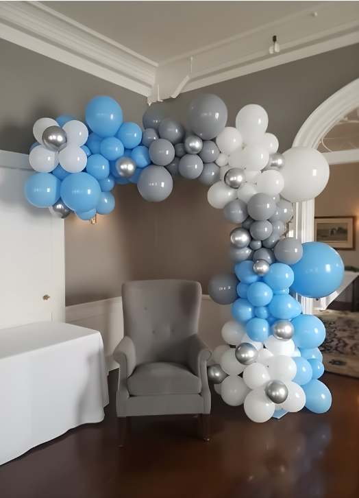 Balloon arch in white, blue, and gray colors over a gray armchair in a room with hardwood floors.