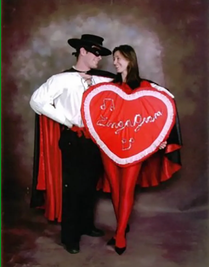 Couple in costumes; man as Zorro, woman in red, holding heart-shaped sign reading 