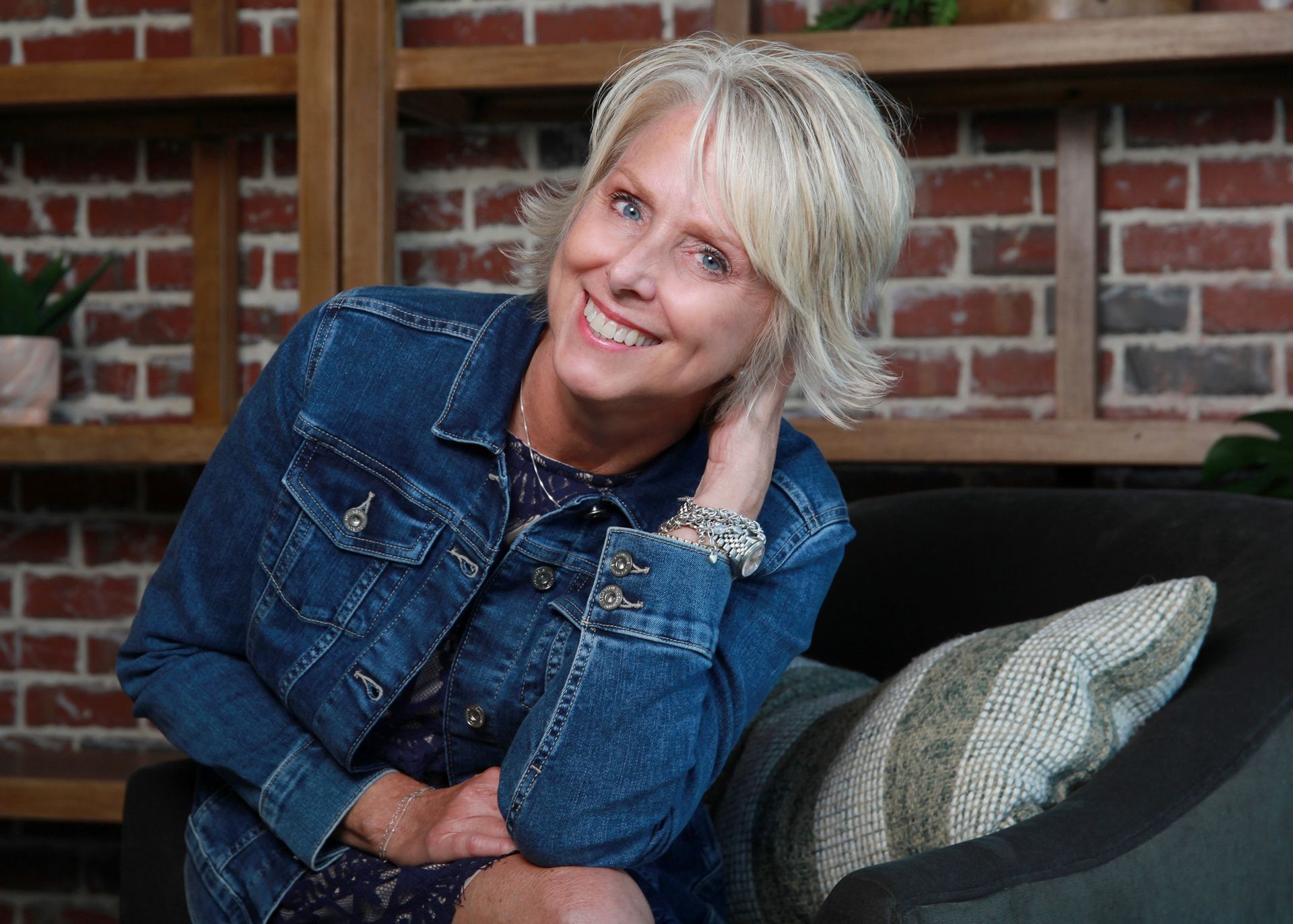 A woman in a denim jacket is sitting on a couch and smiling.