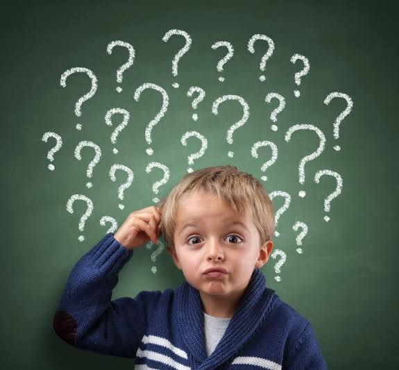 A young boy is scratching his head in front of a chalkboard full of question marks