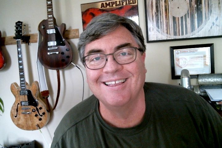 A man wearing glasses is smiling in front of guitars and a poster that says amplified