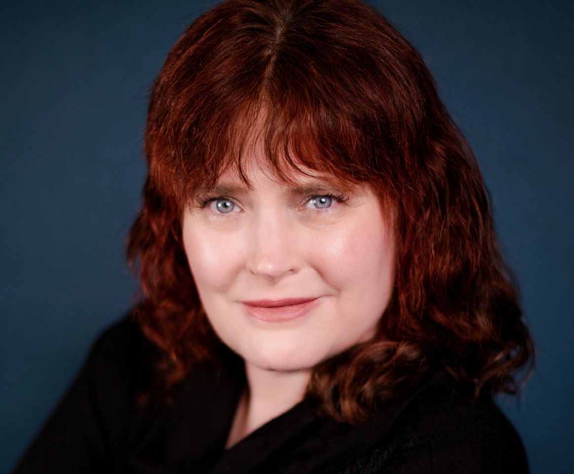 A woman with red hair and blue eyes is smiling for the camera.