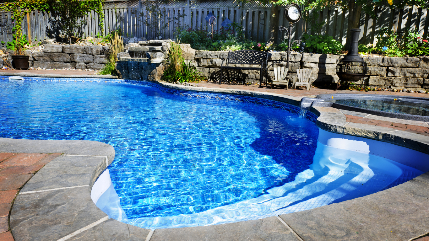 Curved blue swimming pool with stone deck, steps, and patio seating in a sunny backyard