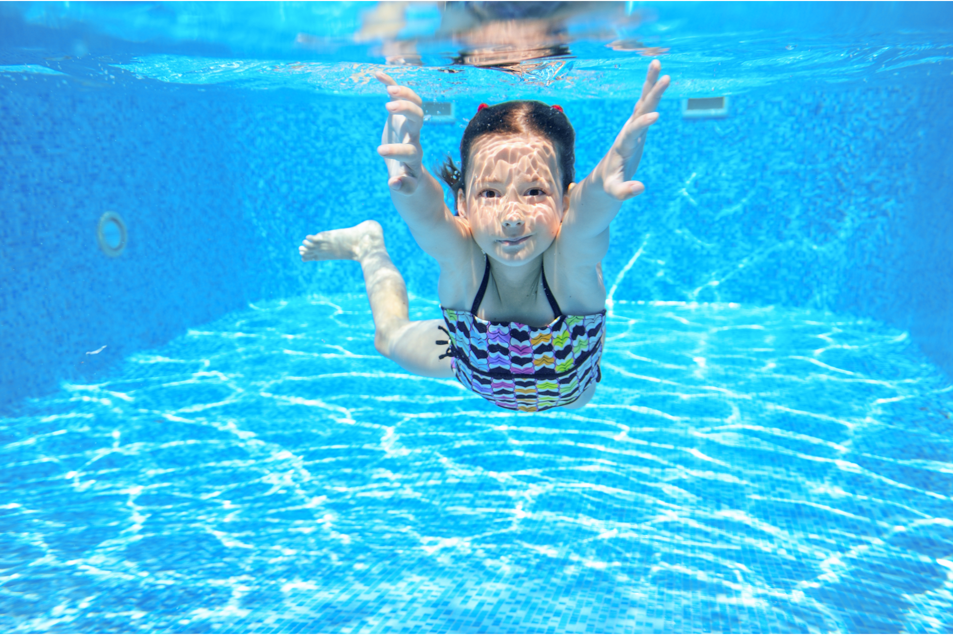Child swimming underwater in a bright blue pool, smiling with arms raised