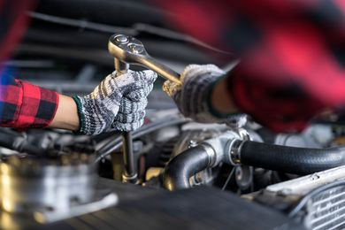 Mechanic using a wrench on a car engine, wearing a plaid shirt and gloves.