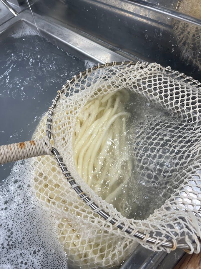 A close-up of cooked udon noodles being lifted out of boiling water with a mesh strainer.
