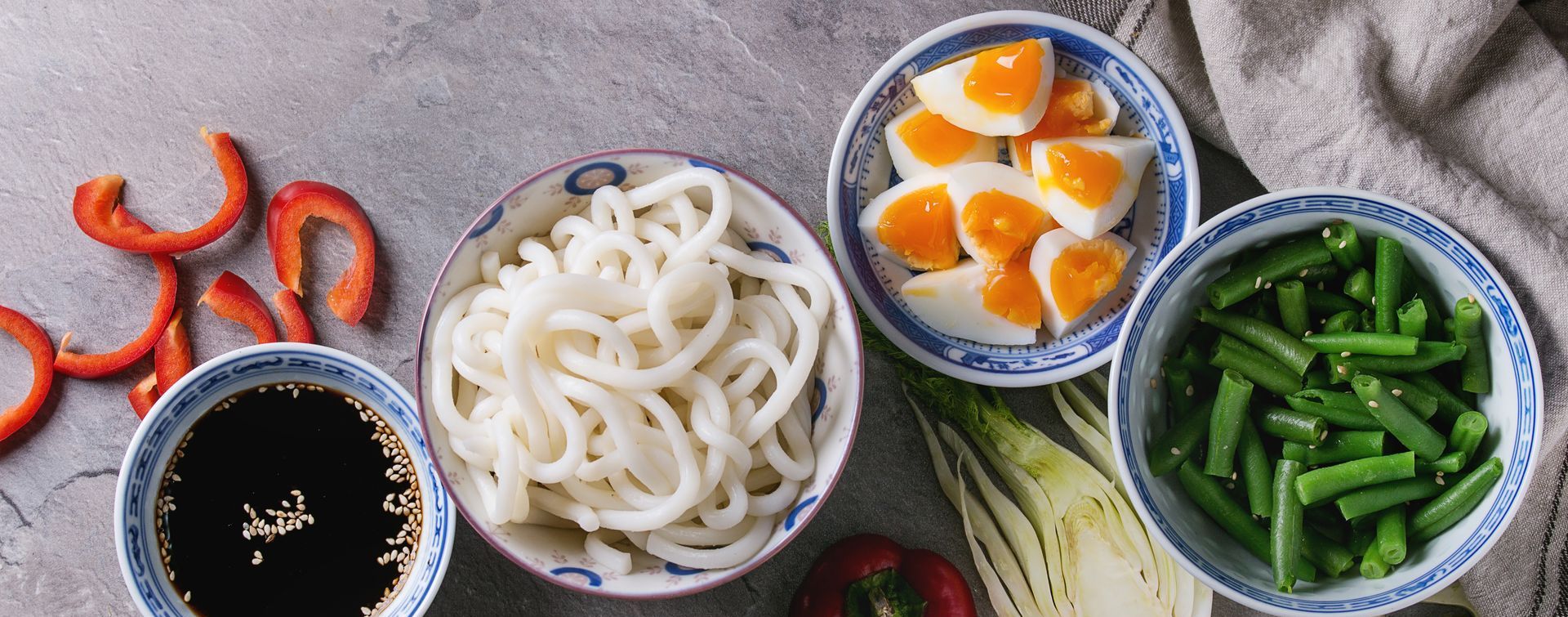 Assorted Japanese side dishes including udon, soft-boiled eggs, green beans, sliced red peppers, and soy sauce with sesame.