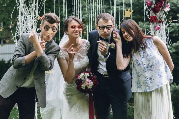 Four adults posing playfully at a wedding, in suits and dresses with floral decorations behind them.