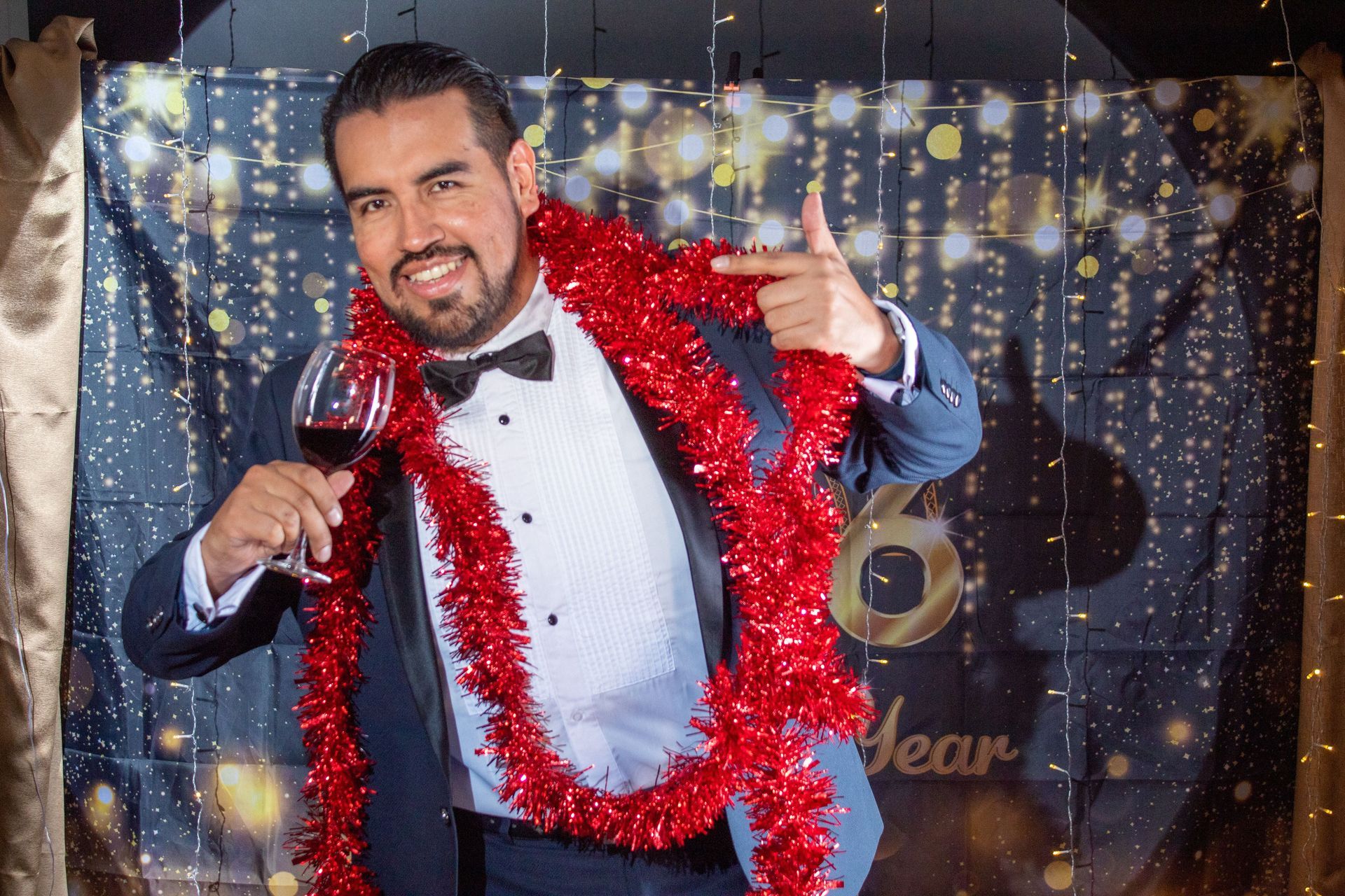 Man smiling in a tuxedo, holding a wine glass and wearing red tinsel garlands at a festive event backdrop