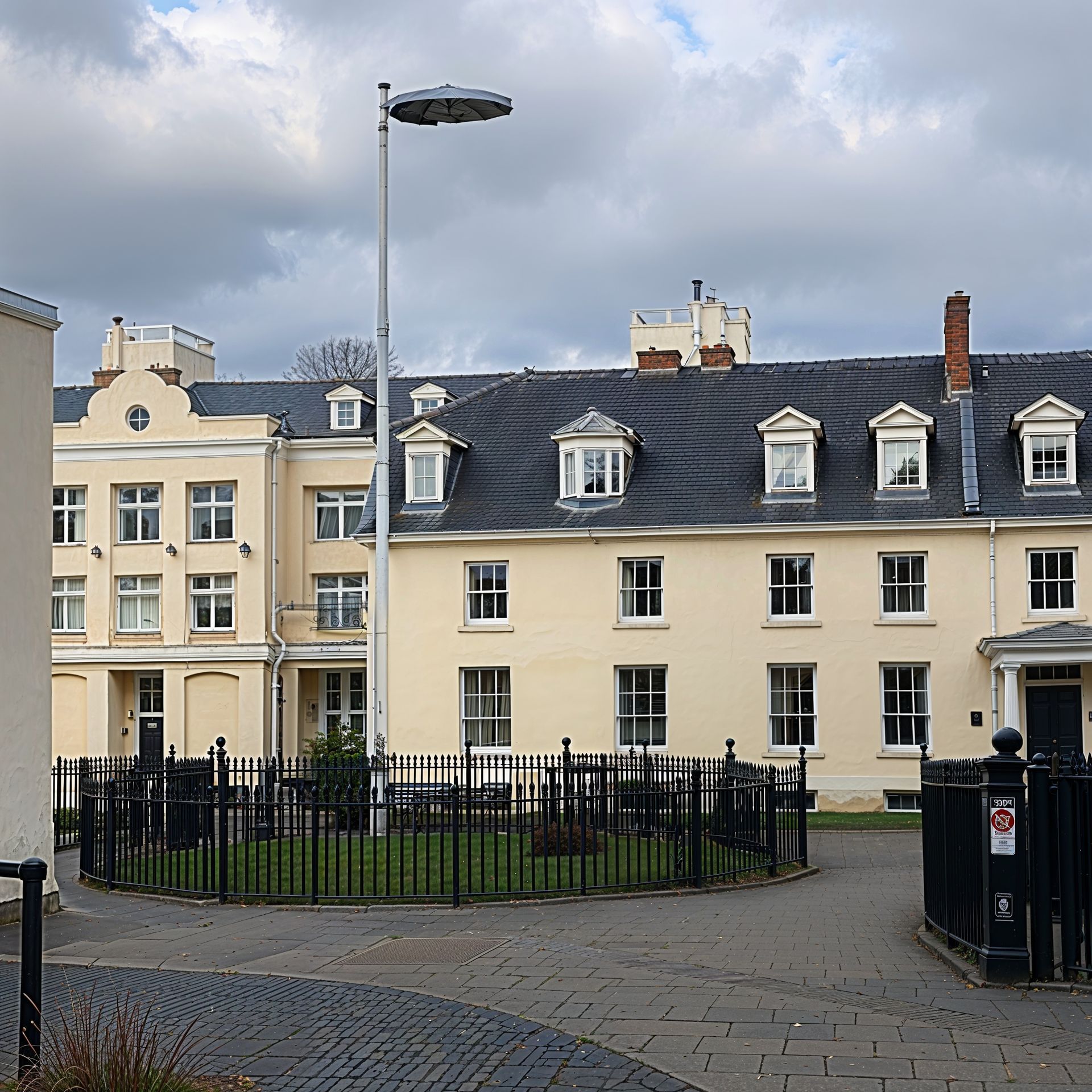 Buildings with light-yellow walls and dark roofs, behind a black fence, in a cobblestone courtyard under cloudy skies.