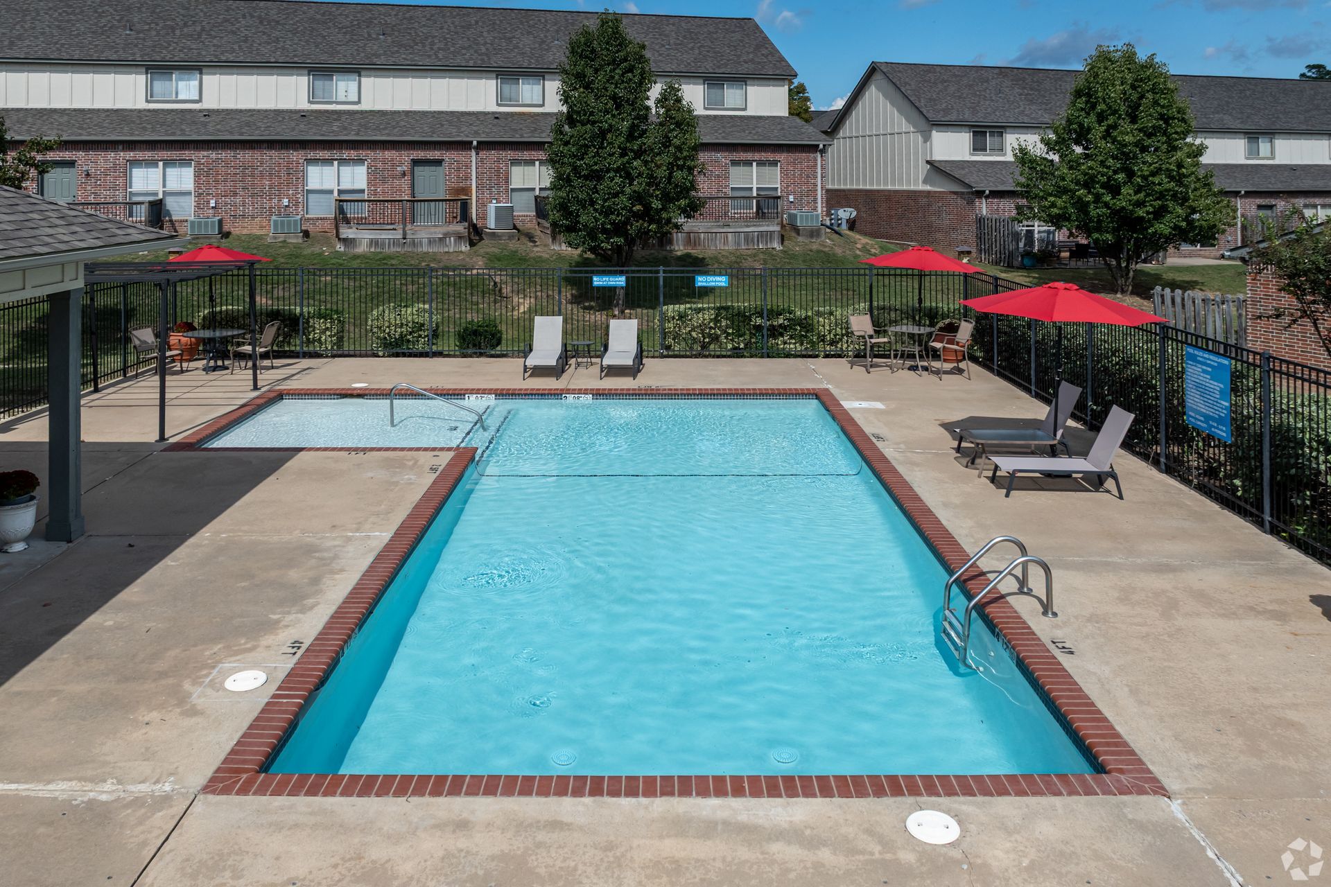Swimming pool surrounded by concrete with red umbrellas and lounge chairs. Apartment buildings in the background.