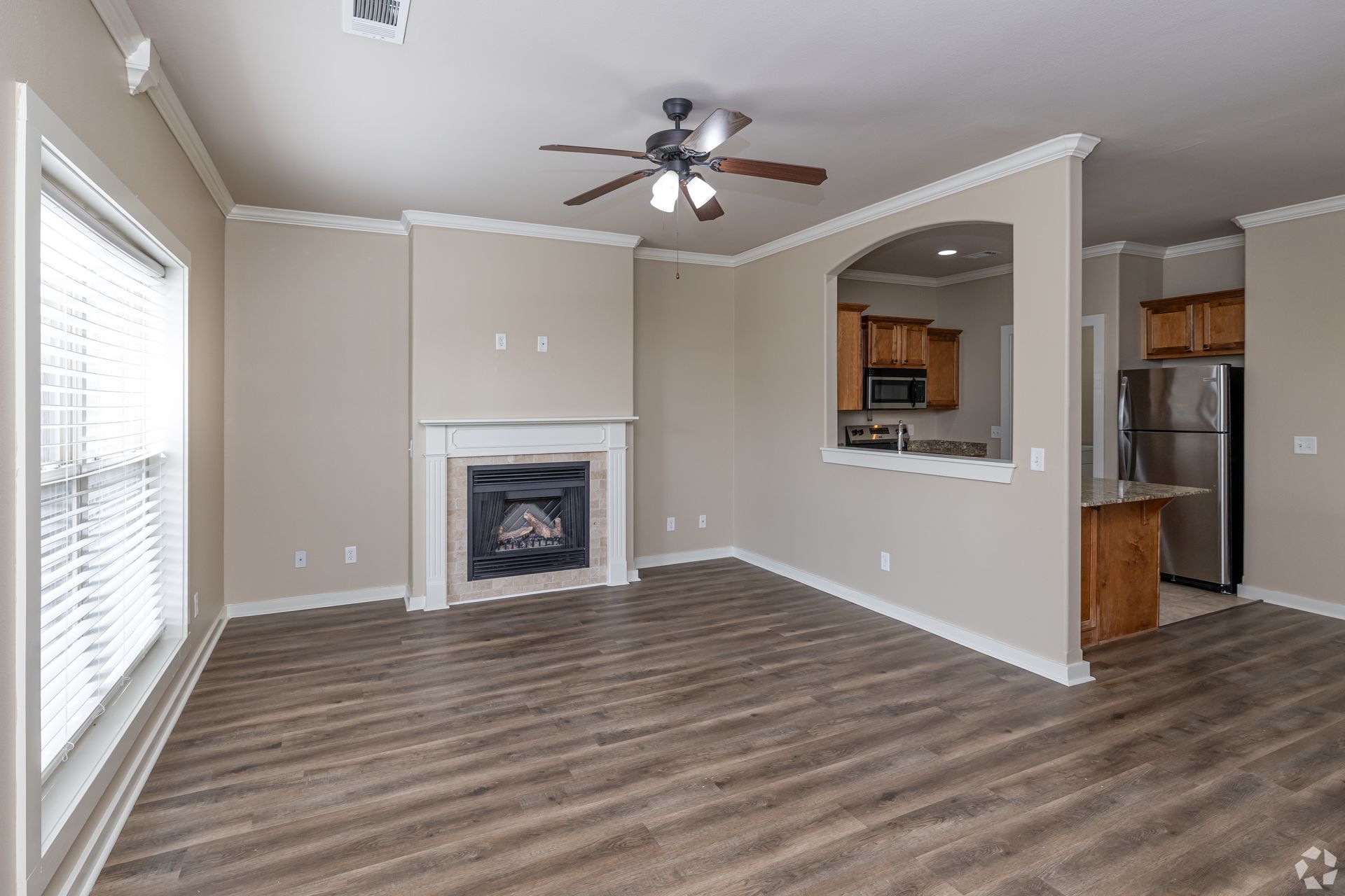 Living room with fireplace, open to kitchen; brown wood flooring, tan walls, white trim.