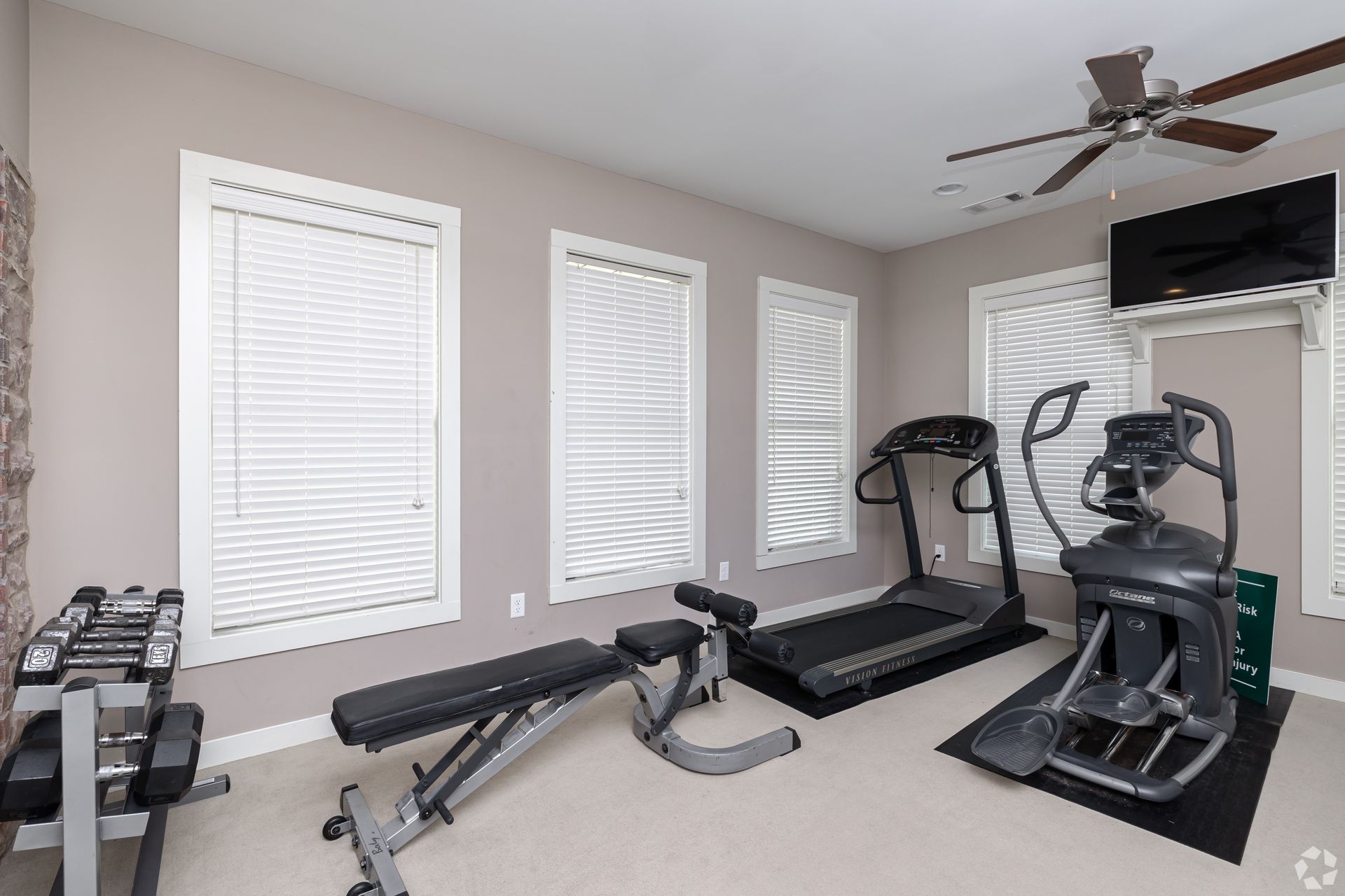 Home gym with exercise equipment: treadmill, elliptical, weight bench, free weights, TV, and three windows.