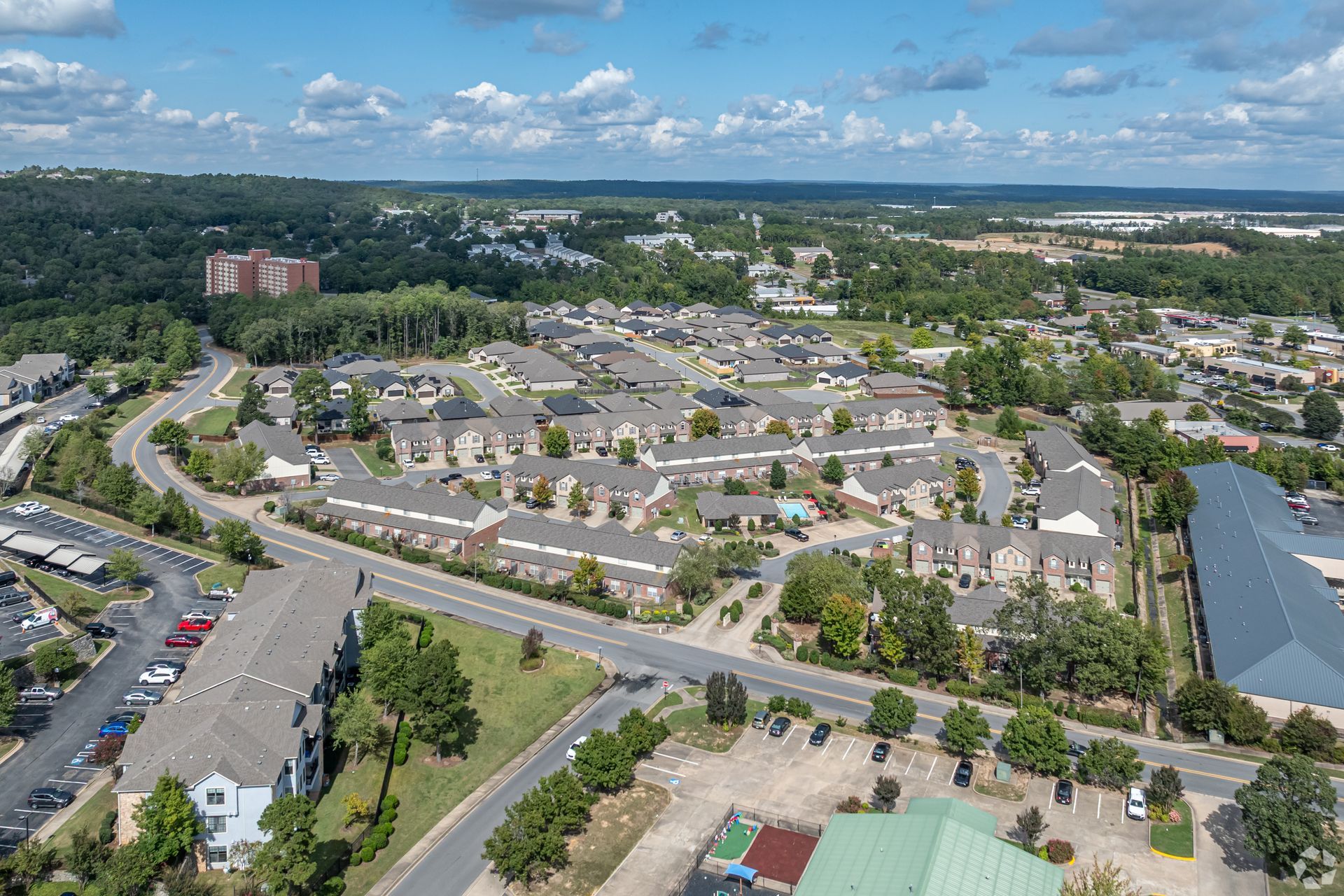 Aerial view of a suburban neighborhood with rows of houses, roads, and trees under a cloudy blue sky.