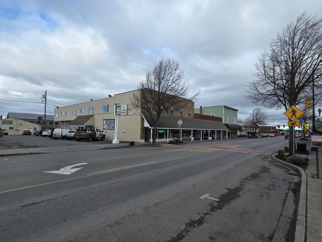 Street scene with commercial buildings and parked cars under a cloudy sky.